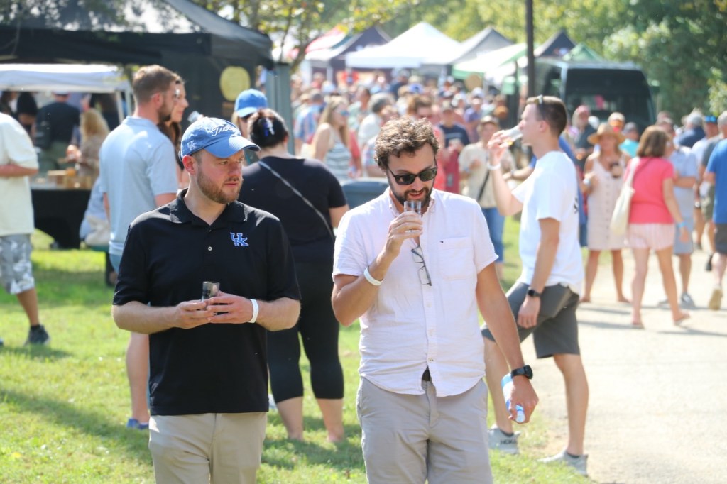Two attendees walk through the Bourbon on the Banks festival grounds holding tasting glasses, with vendor tents and a crowd of people in the background.